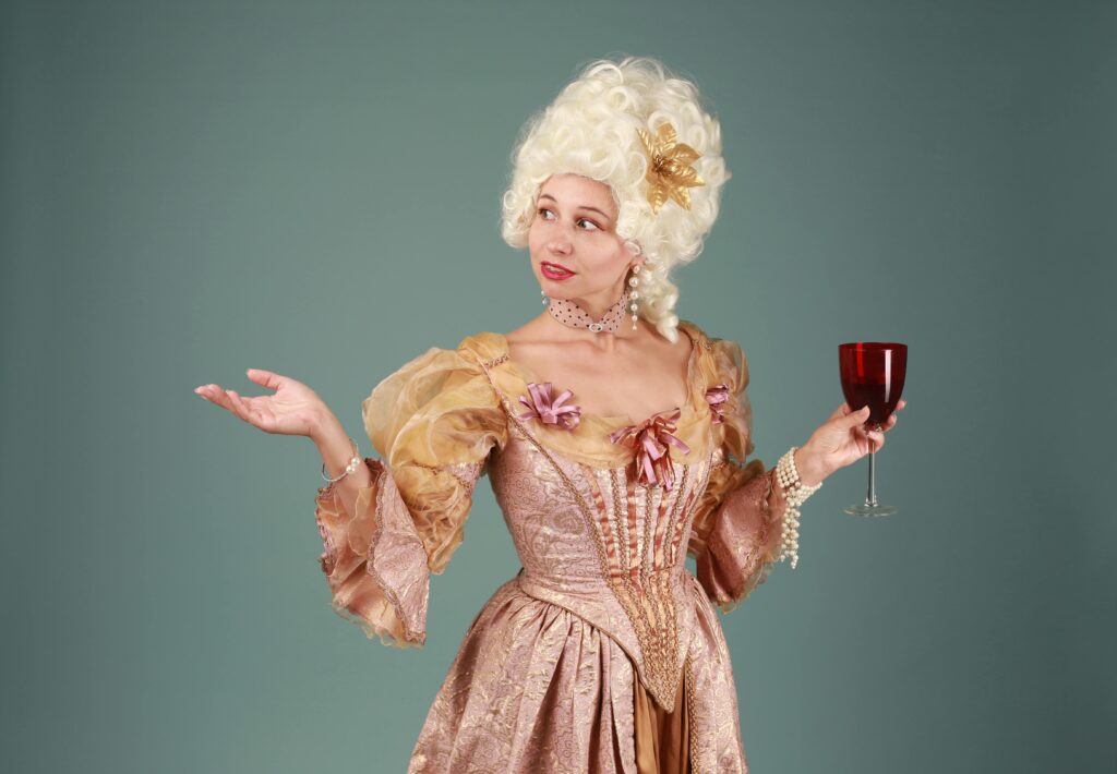 Woman in a Rococo-style dress holding a glass of red wine against a gray background.