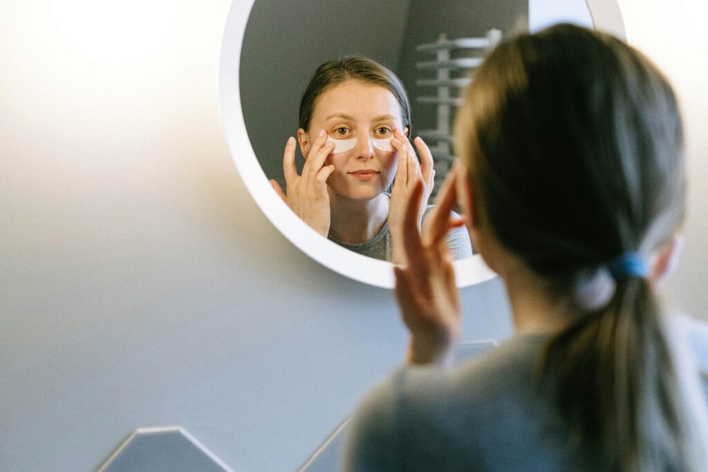 A woman applying under-eye patches while looking in a round wall mirror, focusing on skincare.